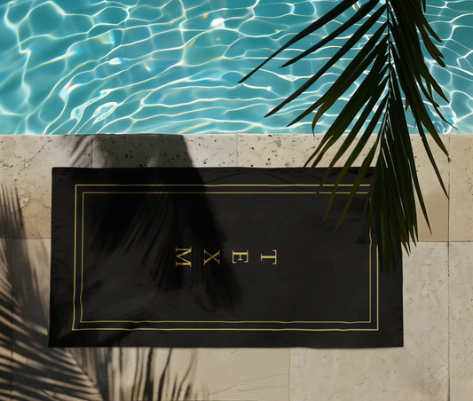 TEXM Ember Black & Gold minimalist beach towel laid on warm sand by the shore